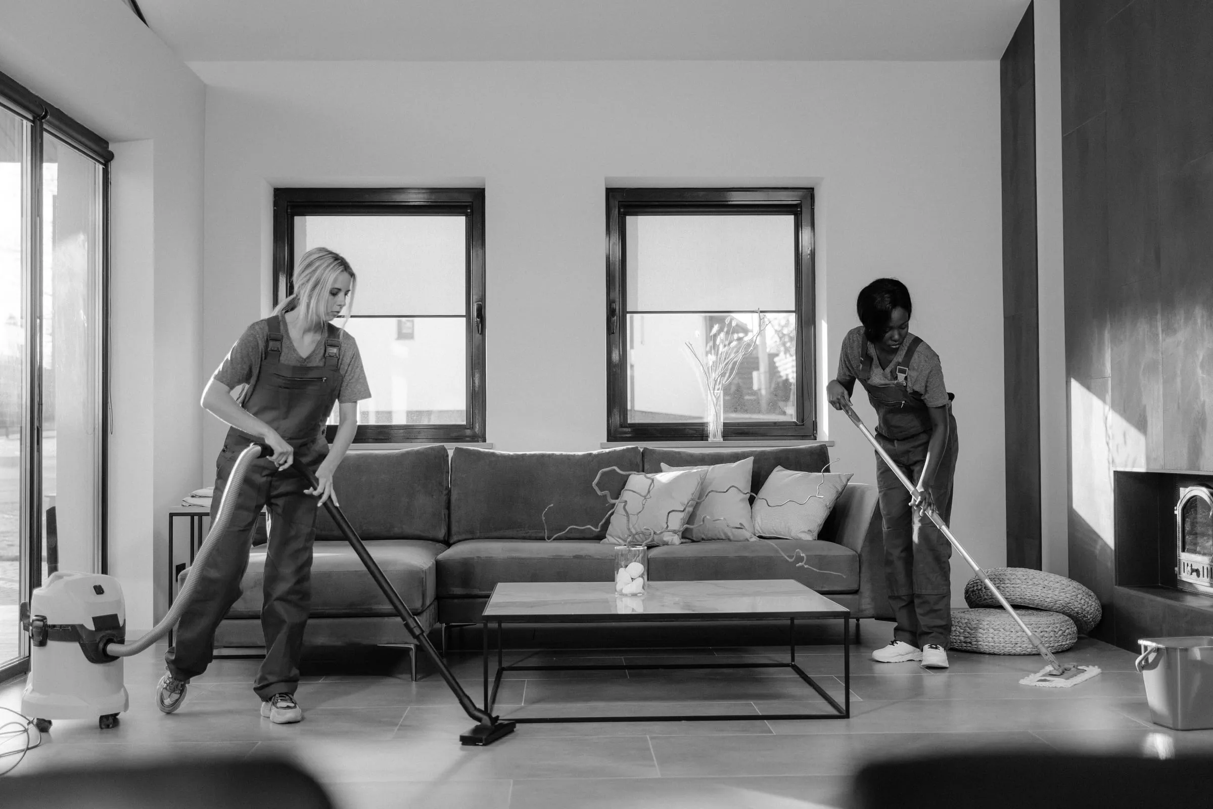 A two-person cleaning crew in work overalls vacuuming and mopping a modern living room